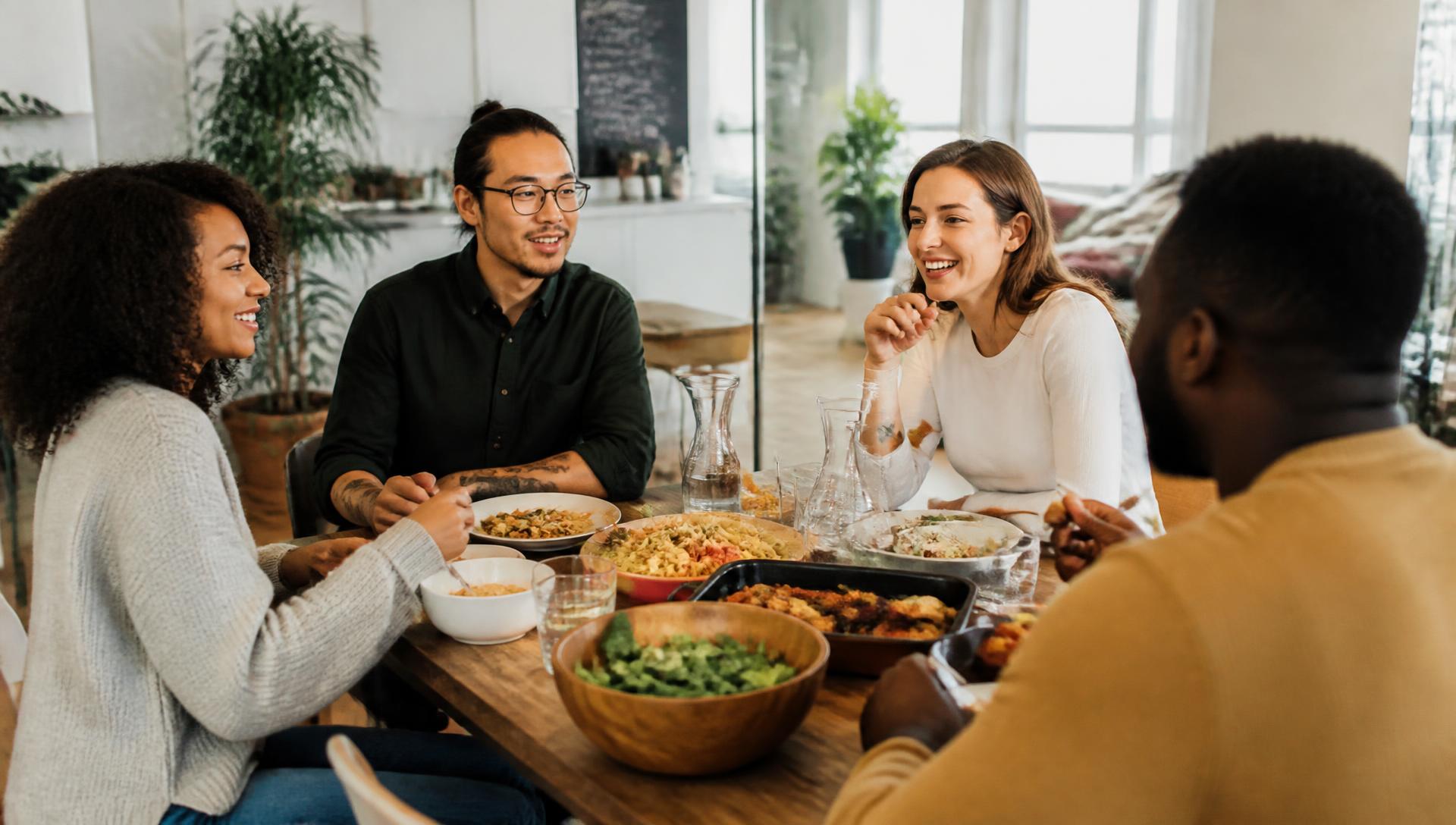 Housemates sharing a relaxed meal and conversation around a wooden dining table in a shared apartment.